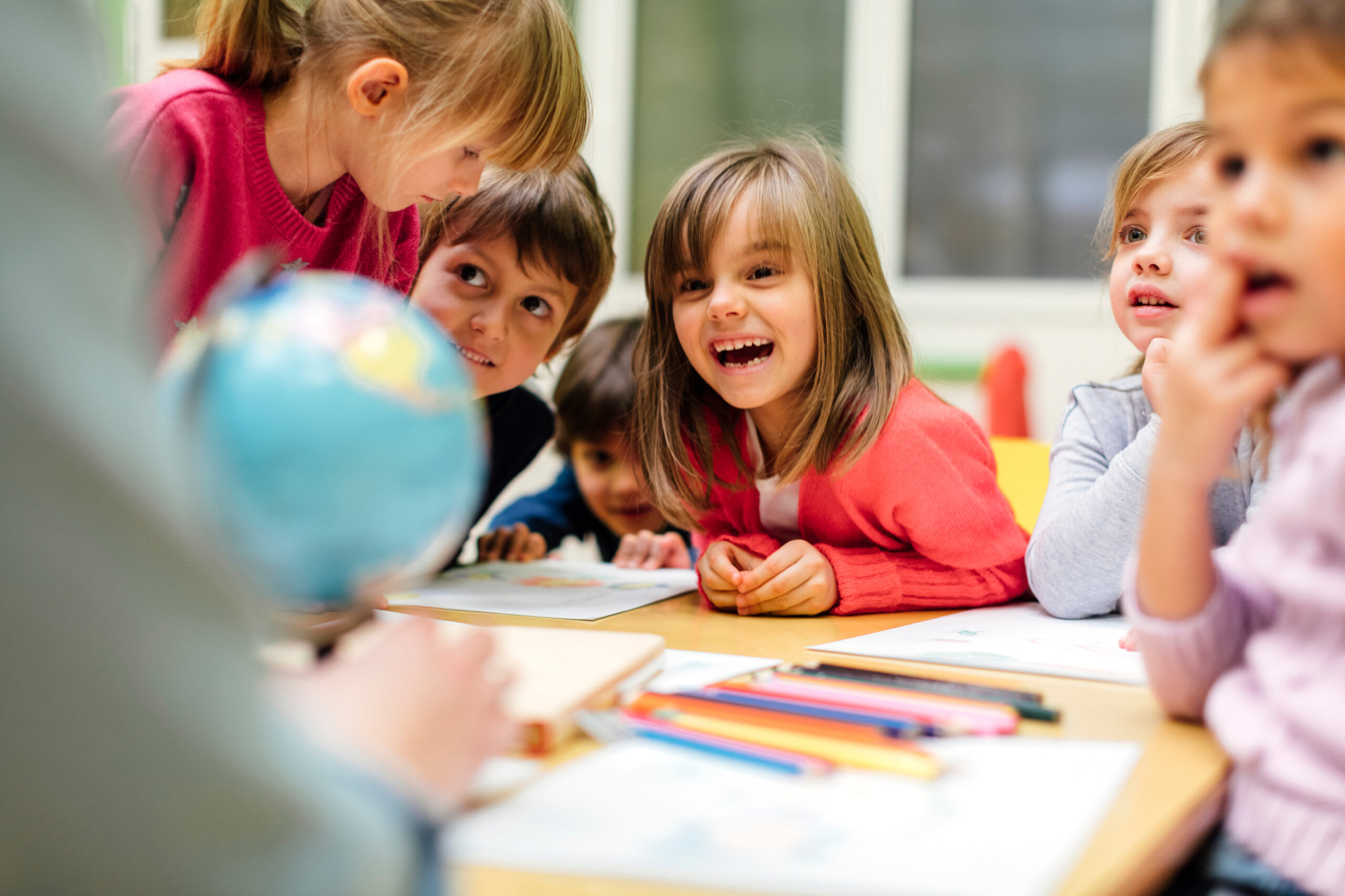 A group of diverse young children happily look at a globe with an adult.